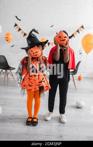 full length of african american kids in halloween costumes covering faces with carved pumpkins at home Stock Photo