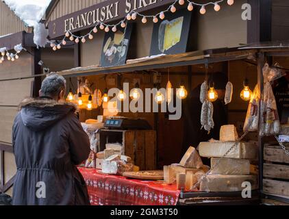 A man standing in front of street holiday market decorated with yellow lights selling cheese and sausages Stock Photo