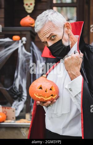 mature man in vampire halloween costume and black medical mask looking at camera while holding carved pumpkin Stock Photo