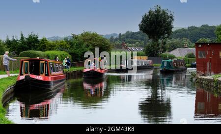 Hired narrowboats moored & sailing (people tying up or casting-off & walkers strolling on towpath) - scenic Leeds Liverpool Canal, Bingley England UK. Stock Photo