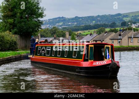 Hired red & green narrowboat sailing & travelling (casting-off) on semi-urban waterway, couple aboard - Leeds Liverpool Canal, Bingley, England, UK. Stock Photo