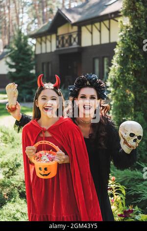 cheerful girl and mother in halloween costumes, with bucket of sweets, skull and toy hand in backyard Stock Photo