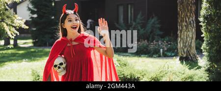 amazed girl in devil halloween costume holding spooky skull and waving hand, banner Stock Photo