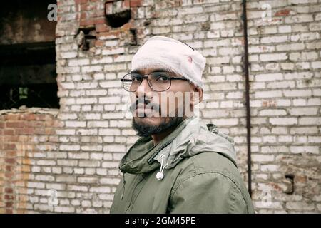 Portrait of miserable young Middle-eastern man with beard having bandaged head, abandoned building in background Stock Photo