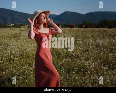 red-haired woman wearing dress and hat on summer evening field of daisies . concept of digital detox, digital cleanse, reconnecting with nature. Stock Photo