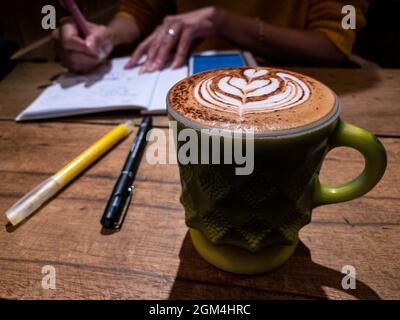 View of woman drinking cappuccino cup with heart shape latte art foam at coffeehouse. Student sitting at wooden table and writing lecture in notebook. Stock Photo
