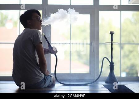 Asian young man in sunglasses smoking a hookah at the window Stock Photo