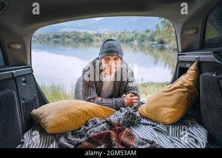 Sincerely smiling middle-aged man dressed in warm knitted clothes and jeans portrait in the cozy car trunk and with beautiful mountain lake view. Warm Stock Photo