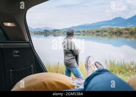 Car trunk view with woman legs in white sneakers on the man dressed warm knitted clothes and jeans enjoying the mountain lake view. Cozy early autumn Stock Photo