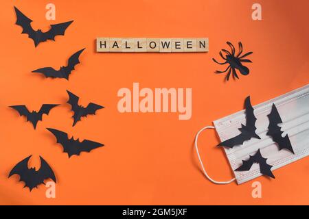 Halloween inscription made from wooden cubes and bats cut from black paper on an orange background with a protective medical mask. Holidays during Stock Photo