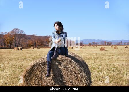 Young brunette woman sitting on top of haystack in field Stock Photo
