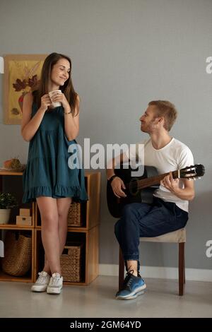Talented creative young man playing guitar for his girlfriend drinking cup of tea or coffee Stock Photo