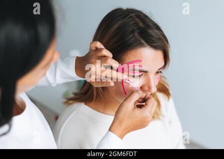 Young beautiful brunette woman with long hair doctor cosmetologist makes face taping for patient in office. Young women do anti-aging procedures with Stock Photo
