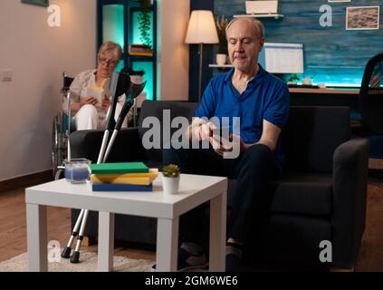 Retired man using smartphone sitting on couch at home having crutches. Disabled elder woman in wheelchair reading book in background. Senior couple with disability in living room Stock Photo