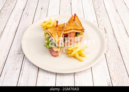club sandwich with chicken, bacon, iceberg lettuce and ham, french fries, red onion, round white plate on white table. Stock Photo