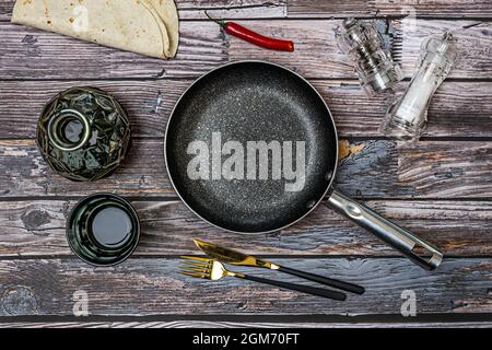 Black frying pan, transparent salt shakers, glass liquor bottle, forks and knives, all on vintage wooden table Stock Photo