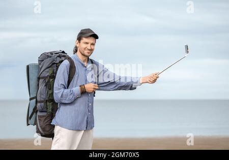 Man, 40 years old, with his tourist backpack and mat is holding a white selfie stick in his hand against a background of a sea on a cloudy day. Stock Photo