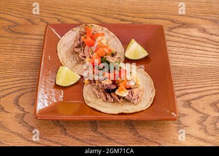 Two tacos of carnitas and vegetables with coriander and a few pieces of lime on an earthenware plate and wooden table Stock Photo