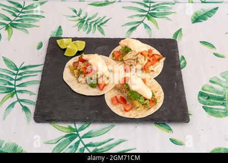 Three tacos in wheat tortillas with a filling of pulled chicken, mayonnaise, pico de gallo, coriander and pieces of lime on a black slate plate Stock Photo