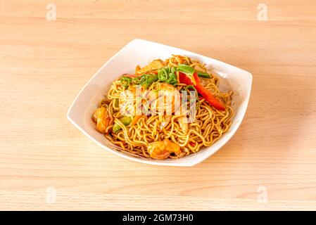 Yakisoba noodles with shrimp and peeled prawns, chopped chives and sauteed peppers and vegetables in white bowl and light wooden table Stock Photo
