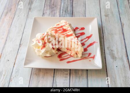 large portion of creamy cheesecake with whipped cream and strawberry syrup on a square white plate Stock Photo