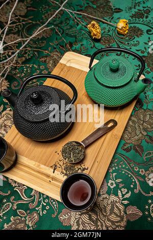 Chinese cast iron teapots in black and one in green with full cups of tea, measuring spoon full of dehydrated tea on a beautiful green cloth Stock Photo
