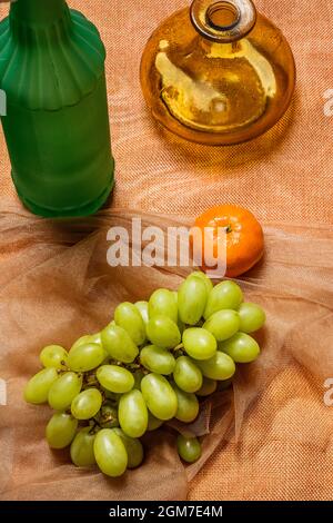 Still life with green grapes, tangerine, green bottle and yellow vase on brown tulle fabric Stock Photo