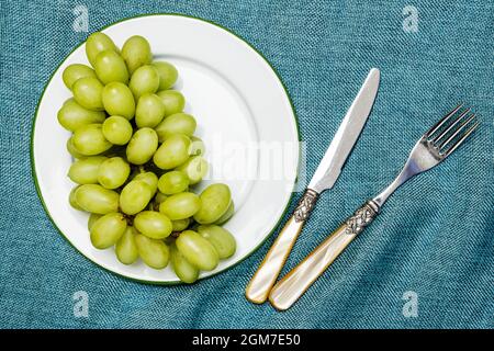 White dessert grapes on enameled metal plate with green rim and vintage cutlery on blue cloth Stock Photo