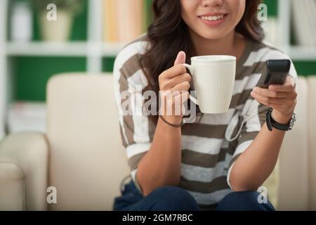Smiling young woman with remote control drinking cup of morning coffee and watching news on tv at home Stock Photo