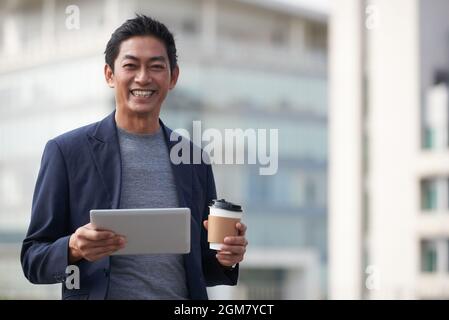 Portrait of happy middle-aged entrepreneur with digital tablet holding cup of take out coffee when standing outdoors Stock Photo