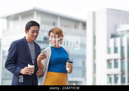 Female entrepreneur showing notification or text message on smartphone to colleague when they are spending time outdoors during coffee break Stock Photo