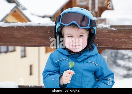 happy cute kid boy wearing helmet, glasses and blue overalls at the ski resort. He eats candy, sits on a bench, rests. Winter active entertainment for Stock Photo