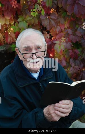cheerful gray-haired old man with glasses is reading the Bible. Portrait of a smiling old man of 88 years on the background of beautiful autumn leaves Stock Photo