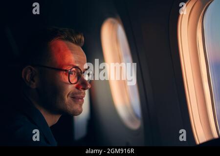 Young man with eyeglasses traveling by airplane. Passenger looking through window during flight at sunset. Stock Photo