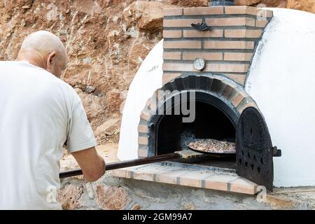 Man putting pizza in handmade white painted wood oven built outdoors with shovel Stock Photo