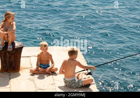 Prvic Sepurine, Croatia - August 25, 2021: Blond children sitting in the sun on the stone pier fishing and looking at the distance, and a sea behind t Stock Photo