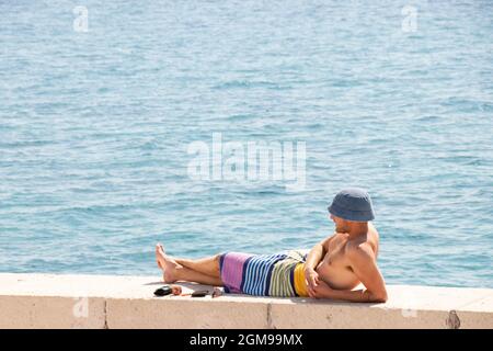 Prvic Sepurine, Croatia - August 25, 2021: Young man wearing hat and wrapped in beach towel lying in the sun on the pier wall looking at the distance, Stock Photo