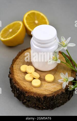 Vitamin C in a white jar on a wooden plate with lemons and flowers. Yellow pills, fruit and herbal supplements. Vertical photo. Side view Stock Photo