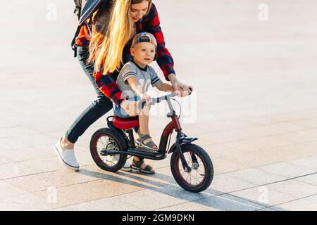 Little happy child learns to ride a bike with her mom in the park in the park, Happy young mom teaches her son to ride a bike in the park on a summer Stock Photo