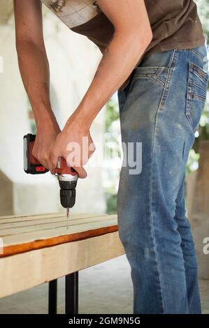 Skilled young male worker is using power screwdriver drilling during construction wooden bench, do it yourself Stock Photo
