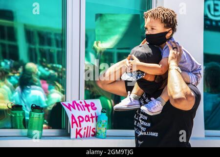 05-30-2020 Tulsa USA Adorable black toddler rides of his fathers shoulders clutching his head, both wearing masks, with reflections of BLM protest blu Stock Photo