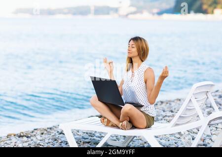 A calm woman sits on a sun lounger near the sea, gets distracted from working at the computer, meditates and removes negative emotions, combines remot Stock Photo
