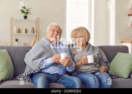 Happy mature couple sitting together on sofa under blanket and holding hot drinks Stock Photo