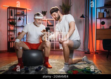 Healthy aged man in activewear sitting on swiss ball and pointing with finger on mobile that holding his son on hands. Middle aged man helping father during domestic workout. Stock Photo