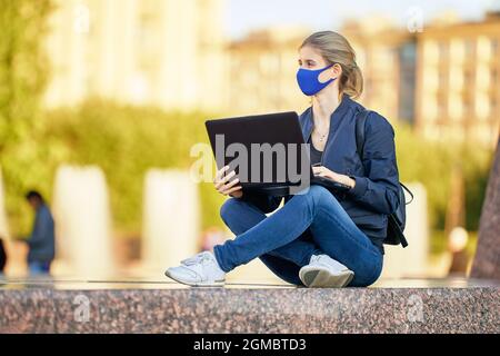 Woman in face mask is making remote work with laptop outdoors. Stock Photo
