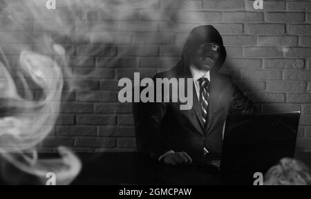 Hacker in black mask and hood at the table in front of the monitor Stock Photo