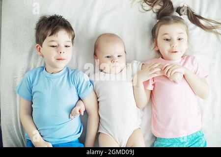 Children lie on the bed next to the newborn baby, little sister. Children's emotions. Stock Photo