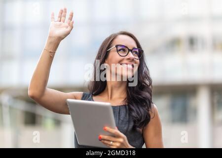 Woman in office clothes and glasses smiles and waves holding a tablet. Stock Photo