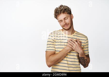 Smiling dreamy young man close his eyes, holds hands on chest and daydreaming, imaging something lovely and heartwarming, cherish memories, white Stock Photo