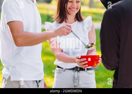 Cropped photo of man pouring the soup into the red bowl and smiling woman holding the bowl, feeding homeless man outdoors, voluntary charity mission. Stock Photo